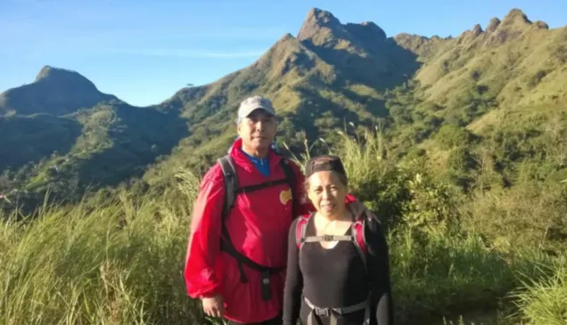 Mariegen and Fons, during a hike to Mount Batulao, pose on a mountain trail with lush green peaks in the background under a clear blue sky.