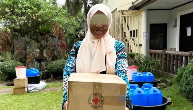 Asniah, smiling, holds an ICRC box, with other donated items in the background.jpg Asniah, smiling, holds an ICRC box, with other donated items in the background.jpg