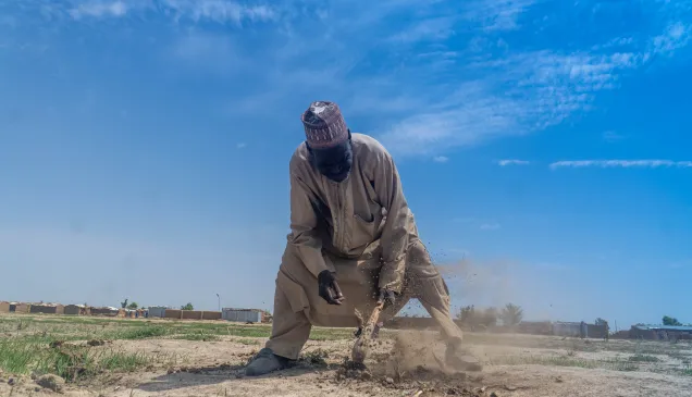 Churi Ibrahim, a 70-year-old farmer, works the dry, dusty soil with a hoe under a bright blue sky in a rural area, with modest shelters visible in the background — a scene reflecting resilience in the face of harsh living conditions.
