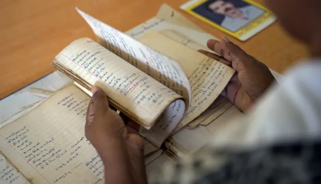 A person flips through the pages of an old, handwritten notebook. Several other aged notebooks lie on the table, along with a photograph of a man placed in the background.