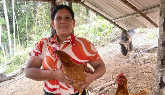 Una mujer, de pie en un gallinero, sostiene con cuidado una gallina entre sus brazos, rodeada de otras aves y jaulas de madera.
