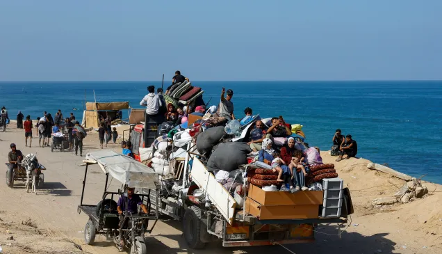 A crowded truck loaded with people and belongings drives along a coastal road, as others walk or travel on carts beside the sea.
