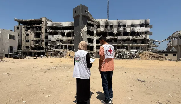 Two humanitarian workers wearing vests with Red Cross and partner organization logos stand in front of a heavily damaged, partially destroyed building, assessing the destruction in Gaza