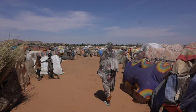 A wide view of a displacement camp in a dry, sandy area, with makeshift shelters covered in tarps and fabric stretching into the distance. Several people walk along the central pathway, including a woman in patterned clothing in the foreground and children on the left. The sky is clear and bright above the crowded camp.