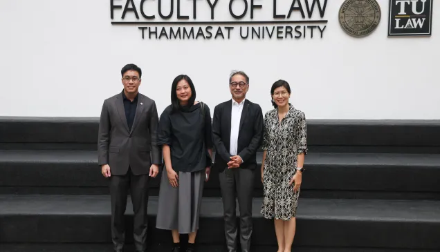 Four people standing and posing for a group photo in front of a wall that reads “Faculty of Law, Thammasat University,” with the university’s emblems displayed above them.
