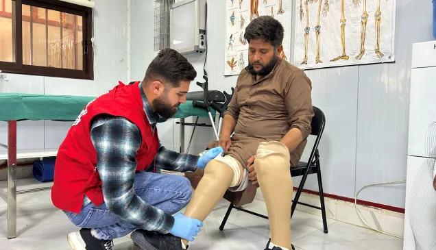 Joumaa, seated on a chair, wears two lower-limb prostheses as an ICRC staff member in a red vest kneels to adjust the fit. They are in a clinical rehabilitation room with anatomical posters on the wall.