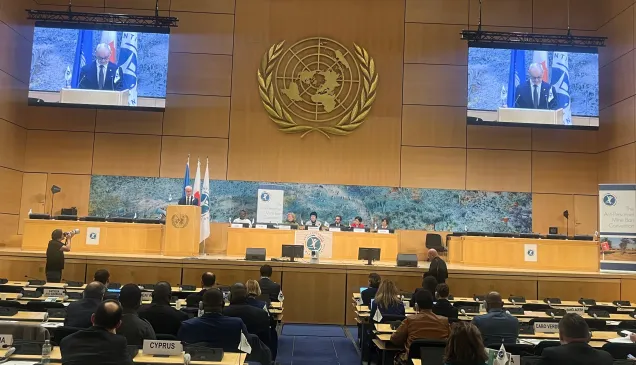 A large conference hall with delegates seated at long rows of desks facing a stage. Two large screens display a speaker at a podium, with the United Nations emblem prominently mounted on the wall behind him. Several panelists sit at a long table on the stage beneath a wide mural. Flags and conference banners are positioned to the side. The audience listens as a photographer captures the scene.