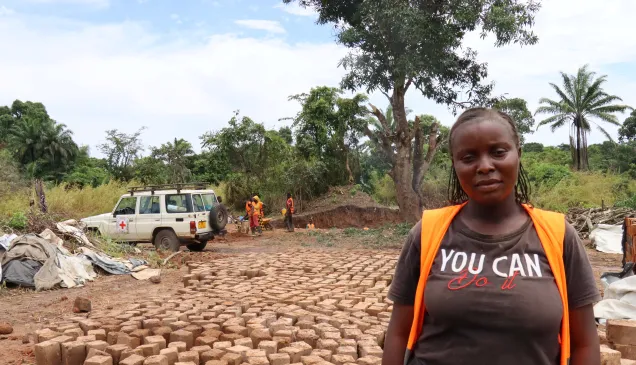 Una mujer mira a la cámara. A sus espaldas hay ladrillos, más personas de la comunidad y un carrod del CICR.