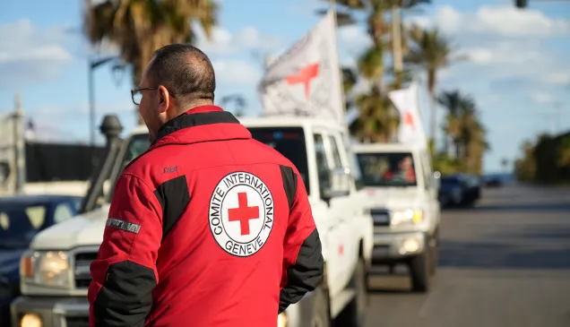 An ICRC staff member wearing a red jacket with the Red Cross emblem stands beside a line of Red Cross vehicles on a coastal road in Lebanon during humanitarian operations amid escalating hostilities.
