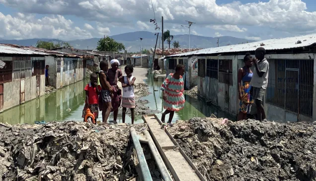 Personas, entre ellas varios niños, se encuentran en un asentamiento con viviendas precarias, donde el agua estancada ha inundado el área. Un estrecho paso improvisado con tablas cruza sobre el agua, mientras las casas de ambos lados muestran condiciones deterioradas bajo un cielo parcialmente nublado.