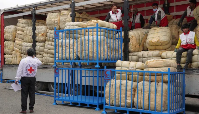 Humanitarian workers unload large bundles of emergency relief supplies from a truck in Iran, with Red Crescent staff coordinating distribution from a loading platform to support people affected by the crisis.