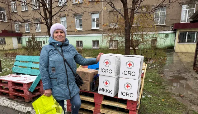 Woman in a blue coat and knit hat stands outdoors beside a stack of white aid boxes marked with red cross symbols on wooden pallets, holding a bright yellow bag; apartment buildings and bare trees are in the background.