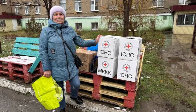 Woman in a blue coat and knit hat stands outdoors beside a stack of white aid boxes marked with red cross symbols on wooden pallets, holding a bright yellow bag; apartment buildings and bare trees are in the background.