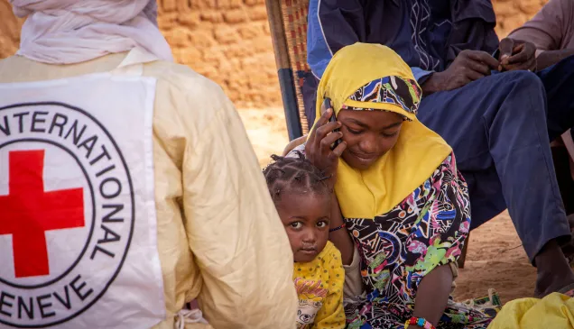 A woman wearing a yellow headscarf sits and speaks on a phone, with a young child beside an ICRC vest in Anderamboukane, Mali, as people gather in the background outdoors.