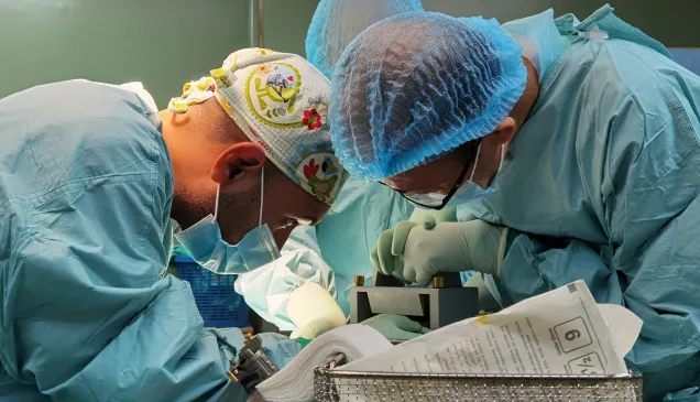 Two surgeons in blue protective gowns and caps perform a delicate operation under bright surgical lights, focusing intently over a patient on the operating table.