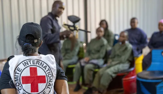 An ICRC staff member oversees a practical session of Law Students during an armed conflict simulation excersise on IHL. She is looking at an instructor holding a mock rifle as students are seated looking attentively looking at the instructor.