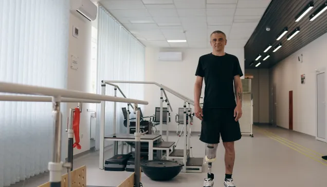 A man with two lower-leg prosthetic limbs stands in a rehabilitation centre beside parallel bars used for gait training. Exercise equipment is visible in the background of the bright therapy room.