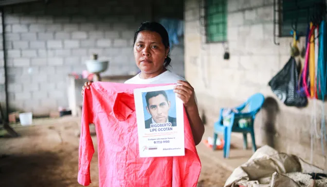 Una mujer sostiene una camisa roja y un cartel de búsqueda con la fotografía de Rigoberto Lipe Ocotán, desaparecido el 13 de junio de 2020. La mujer mira directamente a la cámara desde el interior de su vivienda.