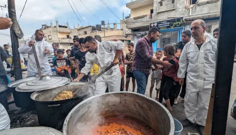 Workers in white overalls stir large vats of food in the community kitchen, people and buildings in the background