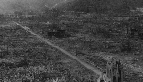 A black and white photograph showing the devastated landscape of Hiroshima after the nuclear bombing in August 1945. The area is flattened and burned, with only a few skeletal buildings and bare trees remaining. A straight dirt road runs through the center, surrounded by rubble and destruction, with mountains visible in the background.