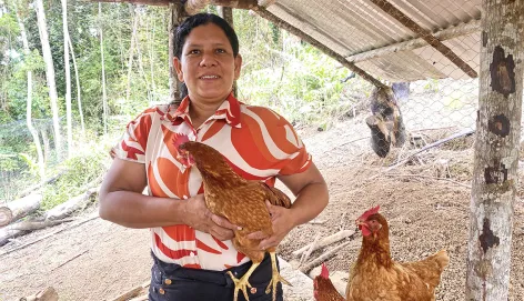 Mujer dentro de un gallinero, sostiene una gallina entre sus manos y sonríe a la cámara.
