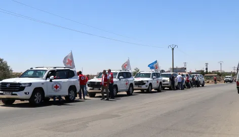 A convoy of white vehicles marked with Red Cross emblems and flags parked on a road under a clear blue sky, with humanitarian workers standing beside them.