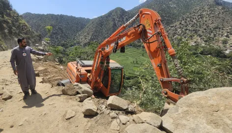 A man wearing a Red Crescent emblem points towards an excavator off a dirt road on a steep hillside in a mountainous area of Afghanistan, following the earthquake.