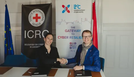 Head of ICRC Global Cyber Hub shaking hands with the CEO of Luxembourg House of Cybersecurity in front of the roll-ups of the respective organizations. With the flags of the European Union and Luxembourg next to the roll-ups.