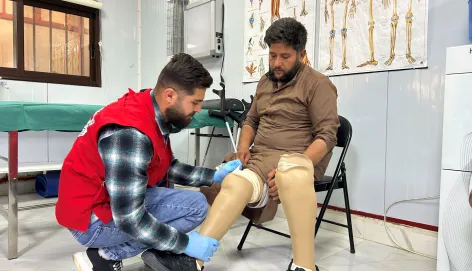 Joumaa, seated on a chair, wears two lower-limb prostheses as an ICRC staff member in a red vest kneels to adjust the fit. They are in a clinical rehabilitation room with anatomical posters on the wall.