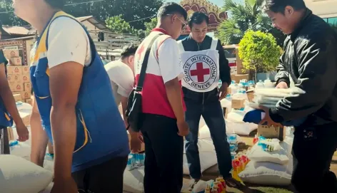 Aid workers distribute relief supplies to people gathered outdoors, with a Red Cross volunteer assisting at a table stacked with bags of goods.