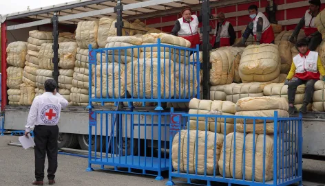Humanitarian workers unload large bundles of emergency relief supplies from a truck in Iran, with Red Crescent staff coordinating distribution from a loading platform to support people affected by the crisis.
