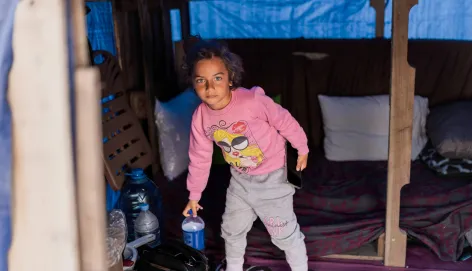 A young girl stands inside a makeshift shelter, holding a small blue container, with bedding and personal belongings arranged around her.