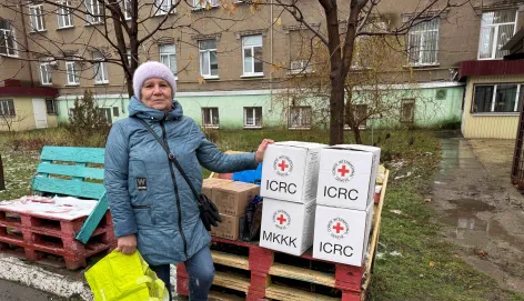 Woman in a blue coat and knit hat stands outdoors beside a stack of white aid boxes marked with red cross symbols on wooden pallets, holding a bright yellow bag; apartment buildings and bare trees are in the background.