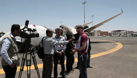 A journalist stands on the tarmac at Sana’a International Airport being interviewed by a local television crew. A passenger aircraft is parked in the background under a clear sky, with airport buildings visible beyond the runway.