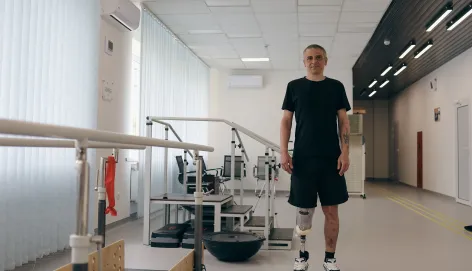 A man with two lower-leg prosthetic limbs stands in a rehabilitation centre beside parallel bars used for gait training. Exercise equipment is visible in the background of the bright therapy room.