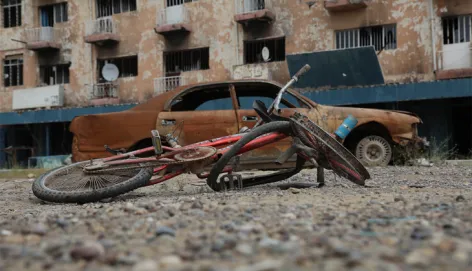 A damaged bicycle lies on a gravel-covered street in front of a burnt-out car and a fire-scorched apartment building, showing destruction in an urban neighbourhood after fighting.