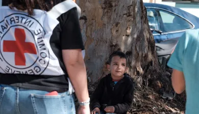 Ghadi sits against a tree and looks up, an ICRC staff member with ICRC vest in the foreground