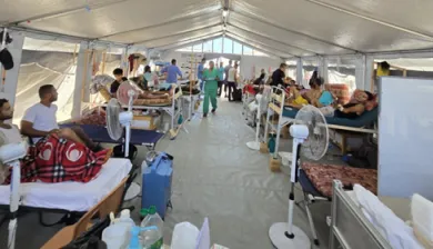 Patients lie on beds inside a crowded tent ward at the Red Cross Field Hospital in Rafah, Gaza. Medical staff in green scrubs move between beds, while fans are placed near patients to provide relief from the heat.