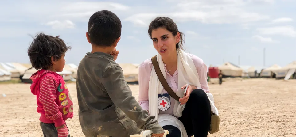 IKRK-Delegierter kniet nieder, um mit zwei kleinen Kindern zu sprechen, von denen eines auf einem Wasserkanister sitzt