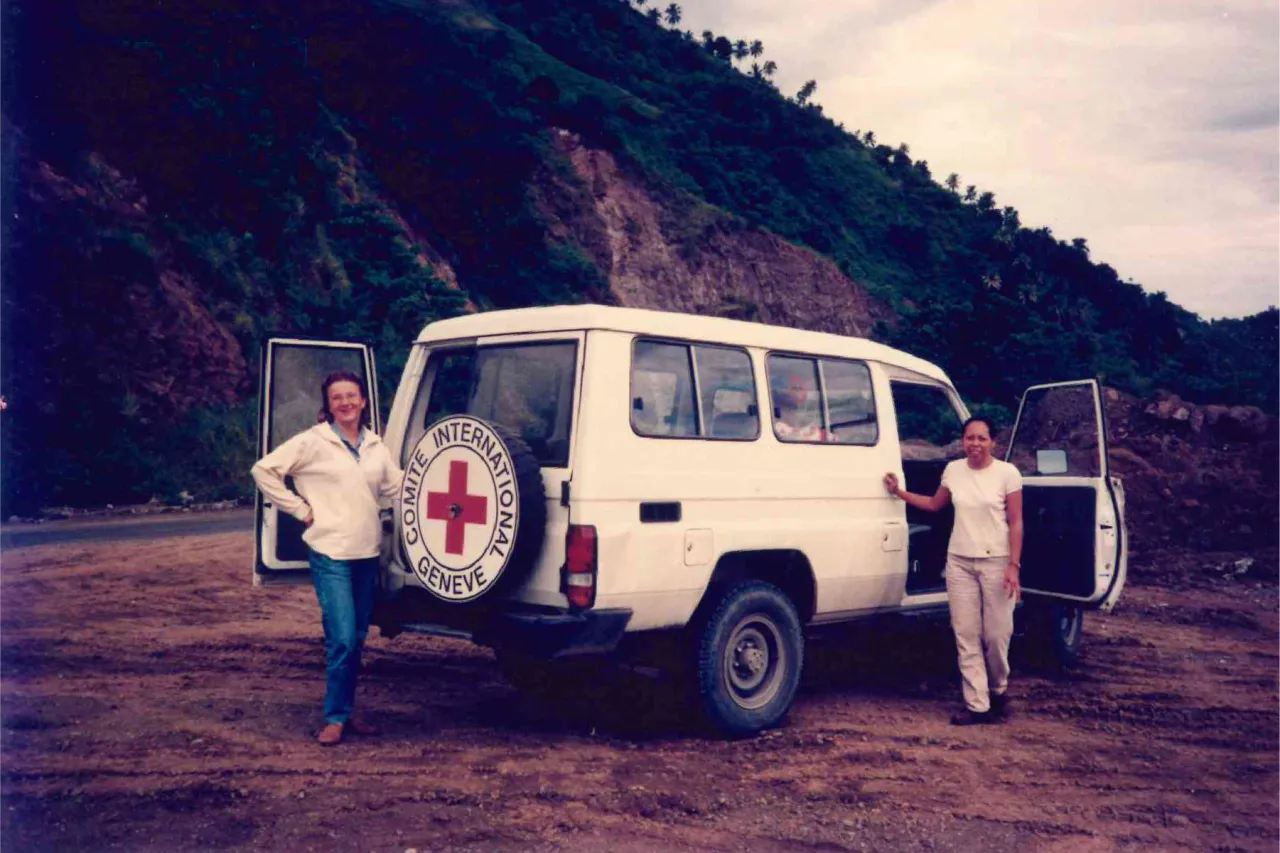 Mariegen and her colleague stand beside a white ICRC vehicle on a dirt road with green hills in the background during a field trip.