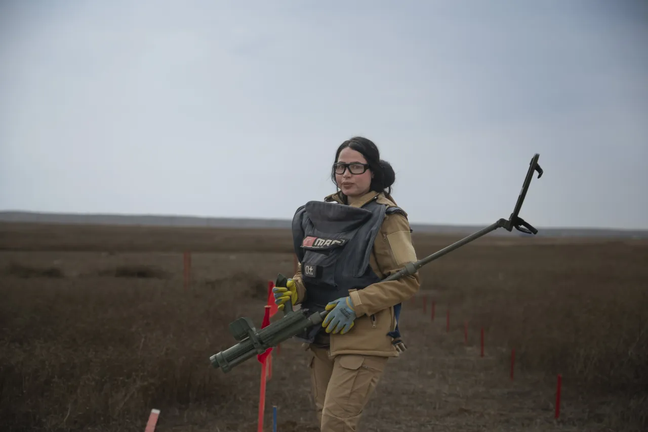 Noora Murad, a female deminer in protective gear, holds equipment while walking through a marked minefield in a barren, open landscape in Iraq.