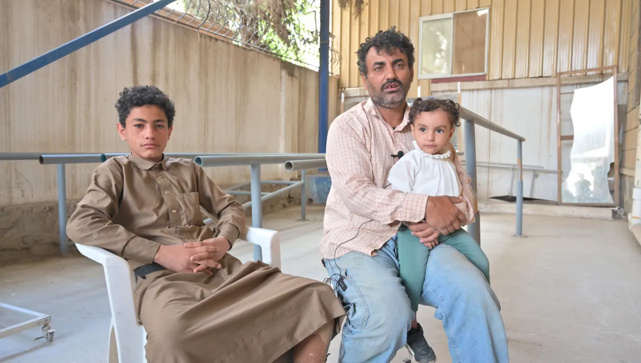 Abdulrahman sits in a chair at an ICRC rehabilitation centre with his father and younger sibling on the chair beside him.
