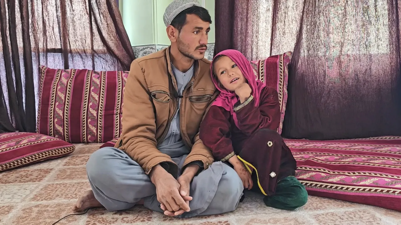 Zahida smiles at the camera, sitting with her father on a rug, with her head on his shoulder.