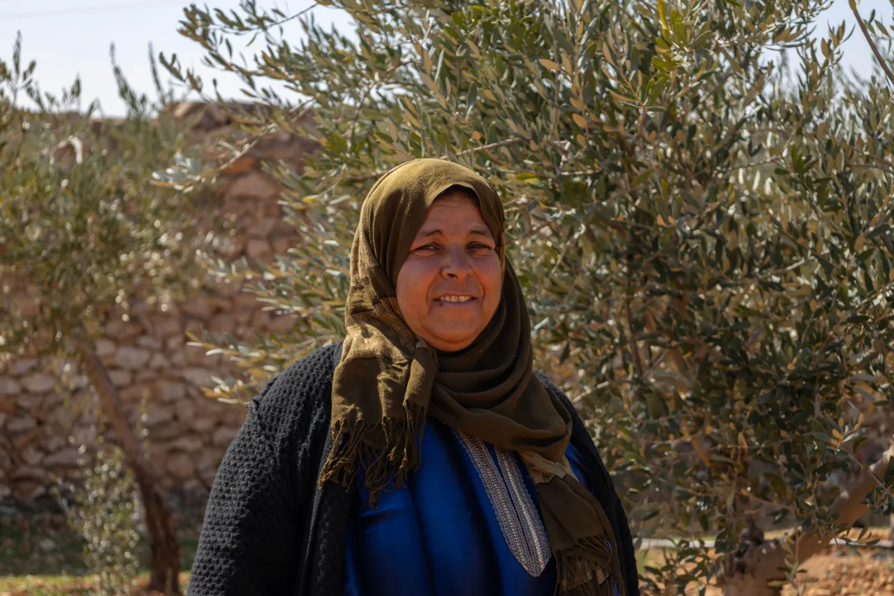 Rabea, a middle-aged woman in a hijab, stands surrounded by olive trees in her village of Barlahin in eastern rural Aleppo, with a gentle smile on her face.