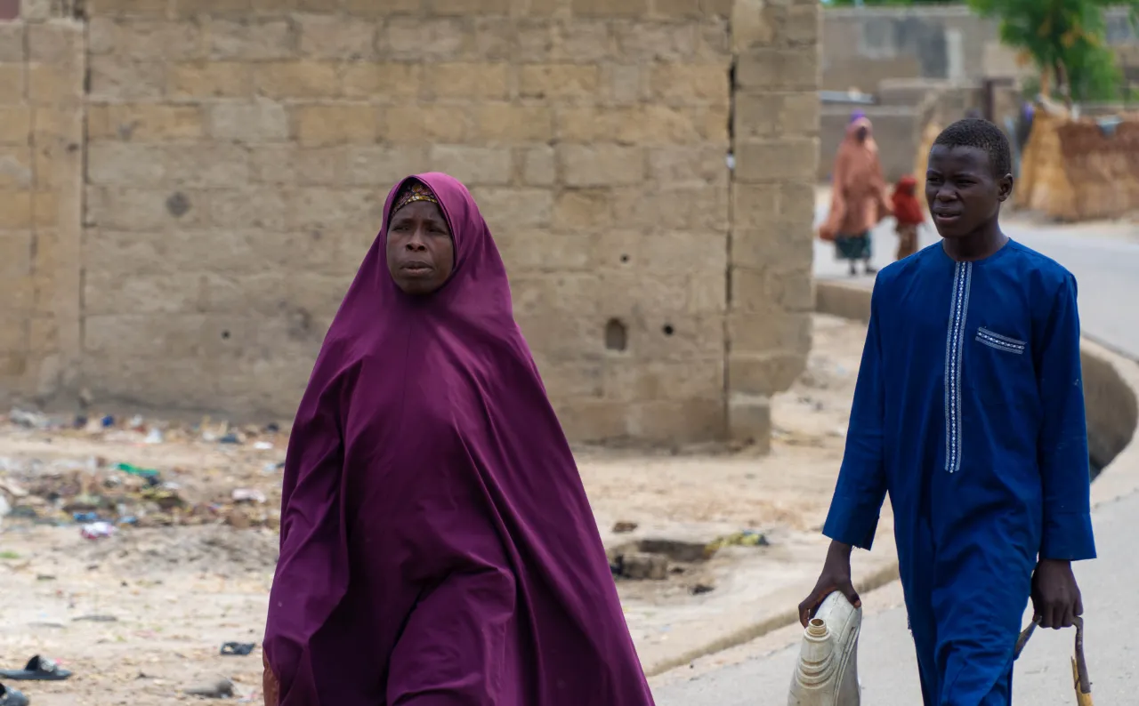 A woman in a deep maroon veil walks alongside a boy in a blue tunic carrying a plastic jerrycan, against a backdrop of modest buildings in a dusty urban setting - a moment of daily life amid challenging conditions.