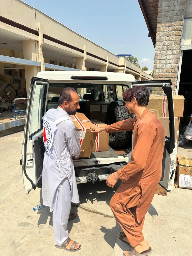 Two men load a box marked with a Red Crescent emblem into the back of a vehicle as part of relief efforts, with stacks of supplies visible in the background.