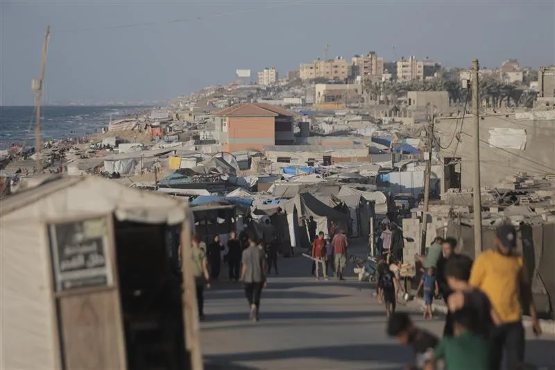 Rows of makeshift tents and shelters crowd a coastal area, with people walking along a narrow road between them, and apartment buildings visible in the background.