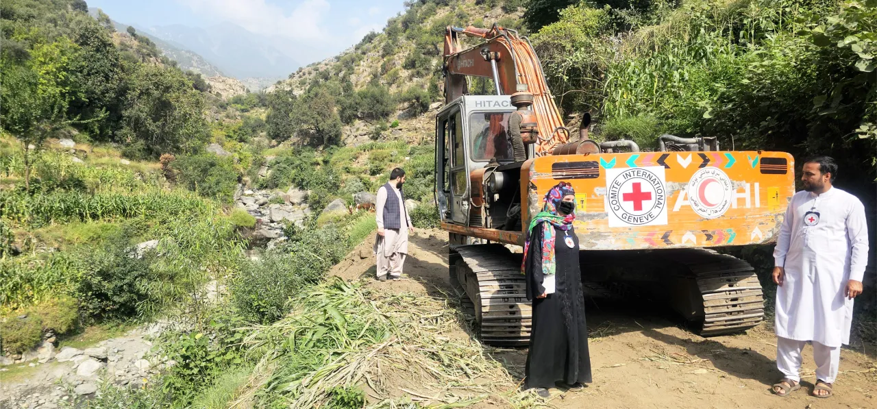 People stand near an excavator marked with Red Cross logos, working on clearing or repairing a rural road in a green, mountainous area.