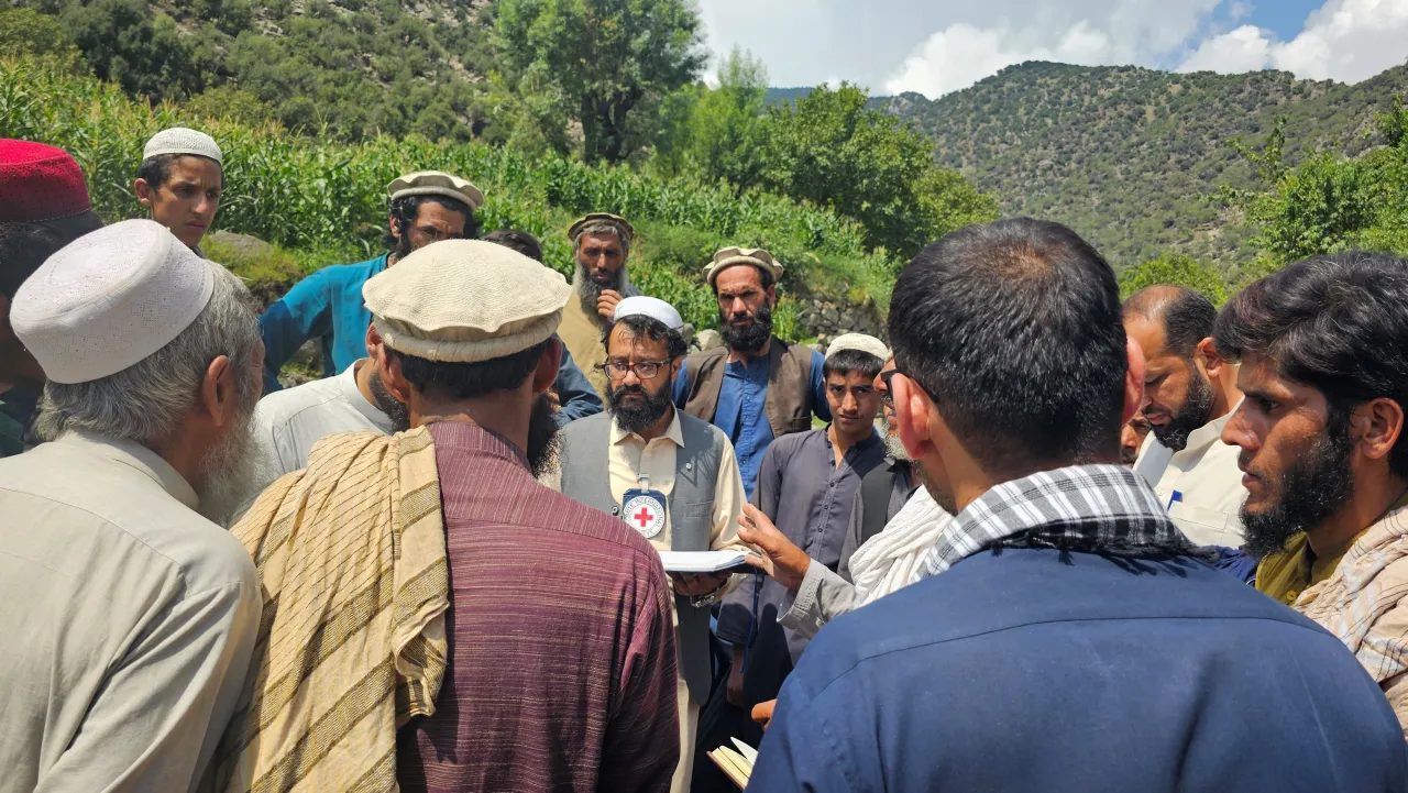 A group of men gather outdoors in a rural, mountainous area, listening to a humanitarian worker wearing a vest with a Red Cross emblem.
