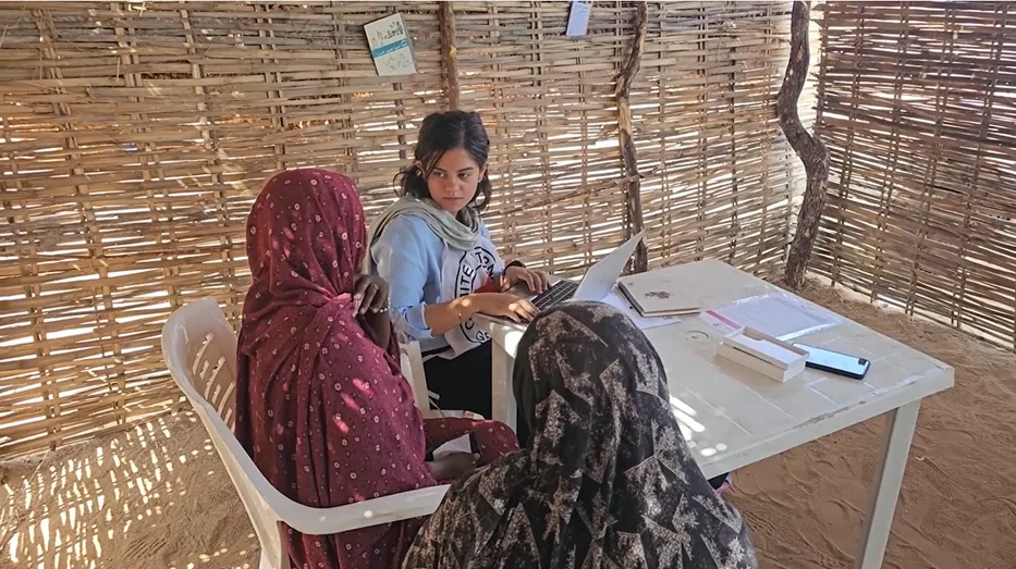 An ICRC worker sits at a small table inside a shelter made of woven branches, speaking with two women who sit opposite her. The women wear patterned headscarves, and the aid worker uses a laptop while taking notes. Sunlight filters through the gaps in the shelter walls.